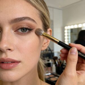 Makeup artist applying eyeshadow with a blending brush to young woman with blonde hair and natural glam makeup look