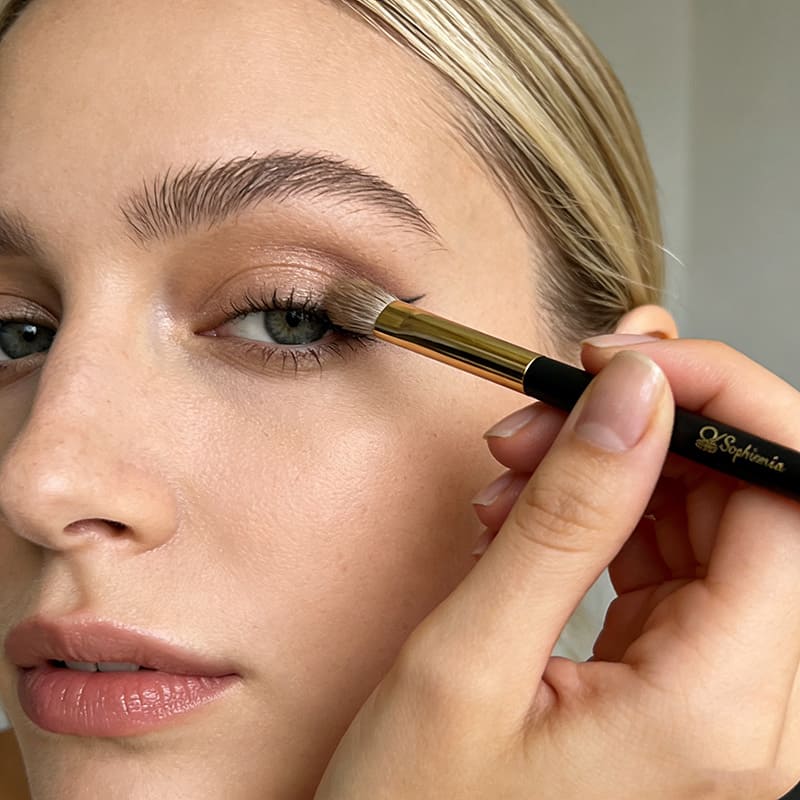 Woman applying eyeshadow with Sophenia makeup brush to eyelid, demonstrating natural eye makeup application technique