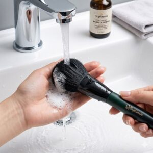 Hands washing a Sophienia makeup brush under running water with foam in a white sink, brush cleanser bottle in background