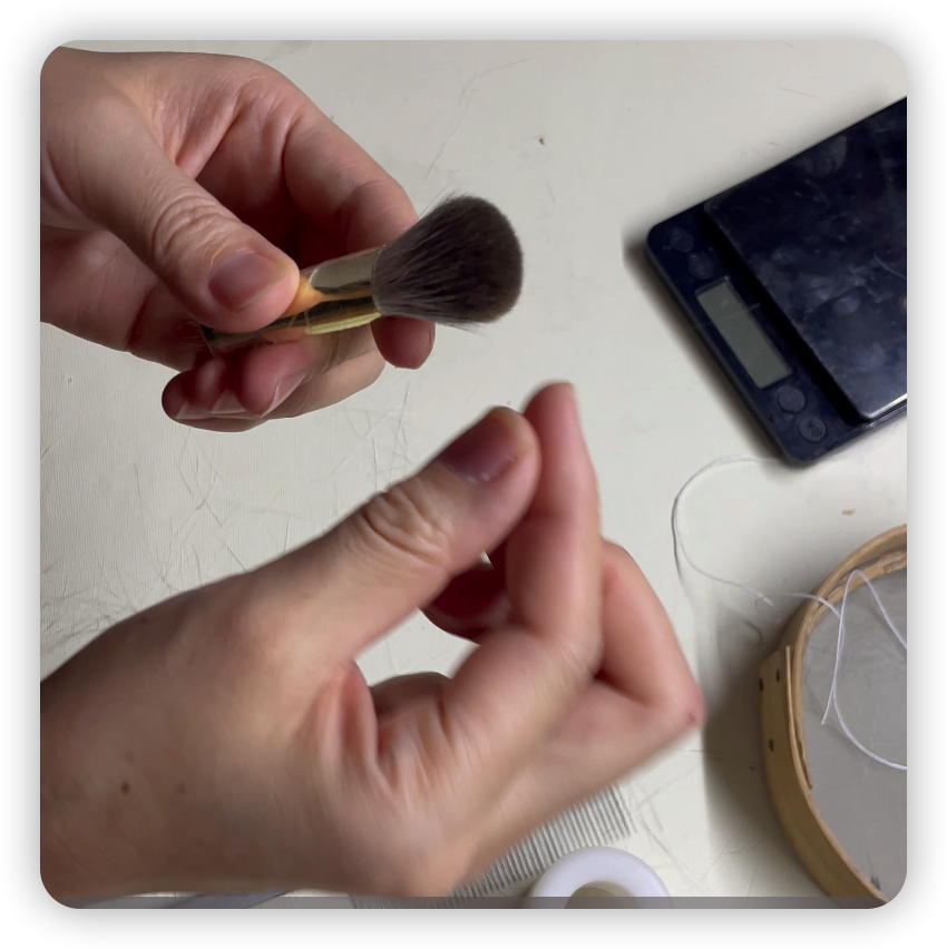 Hands holding a small makeup powder brush over a white surface with digital scale and embroidery hoop nearby